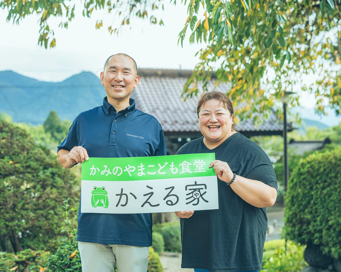こども食堂の看板を持ち笑顔でこちらに顔をむける男性と女性